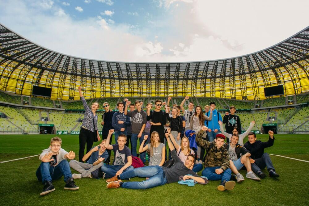 Zdjęcie przedstawia wnętrze stadionu. Na jego tle stoi grupa szęśliwej młodzieży.