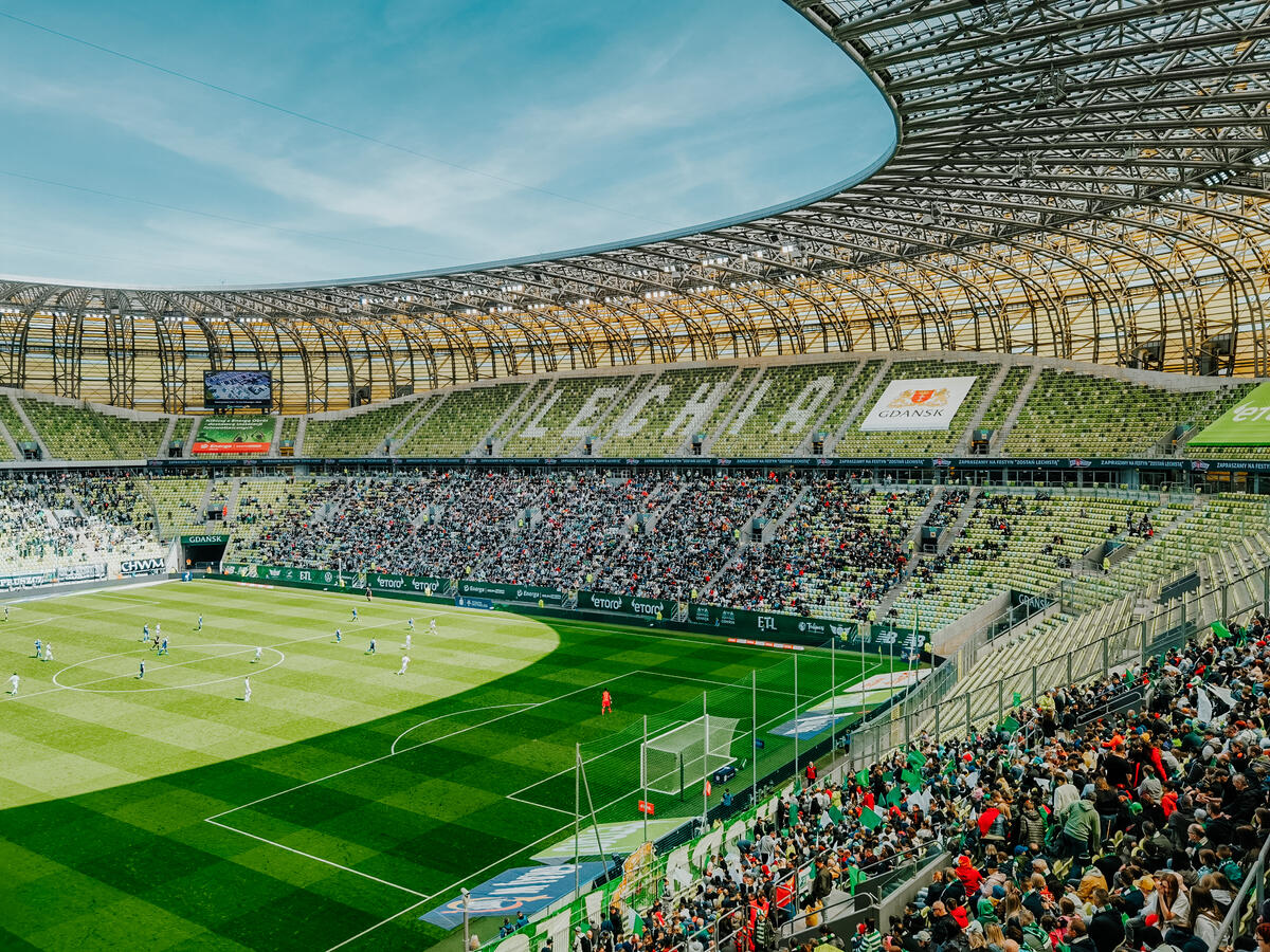 Zdjęcie stadionu wewnątrz przedstawiające kibiców zasiadających na trybunach, którzy oglądają mecz.